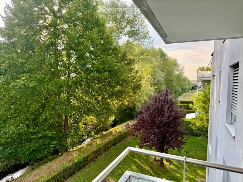 Balcony overlooking a green, tree-lined area with a small garden and neighboring buildings.