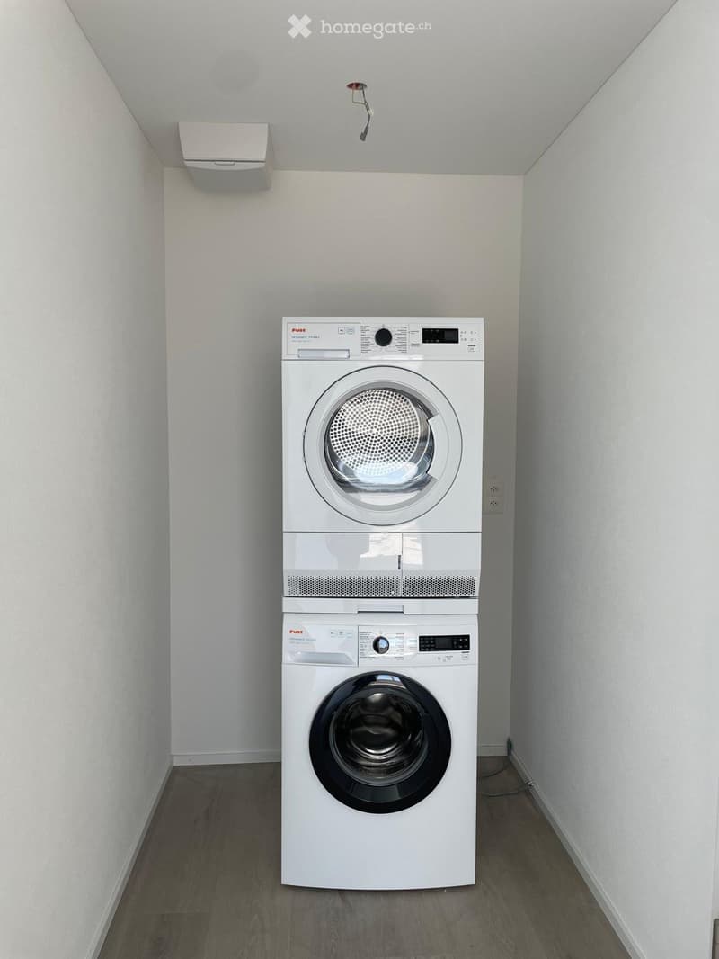 A small, well-lit room with a stacked washer and dryer unit against a white wall, featuring a wooden floor and a light switch on the wall.