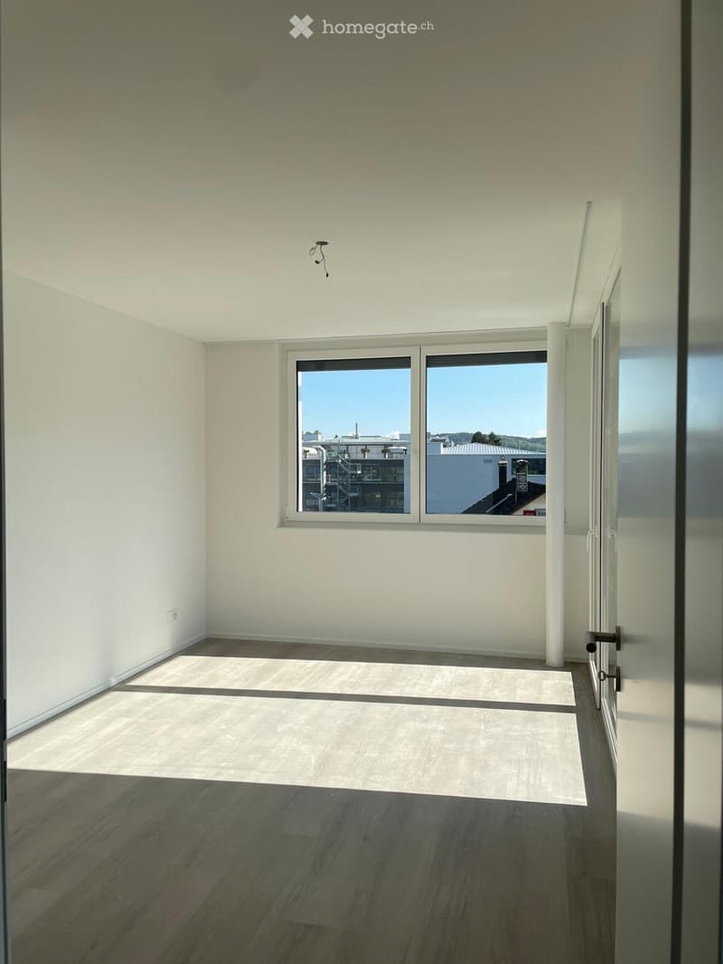 Empty room with light-colored walls, wooden flooring, and large windows offering a view of the outside.