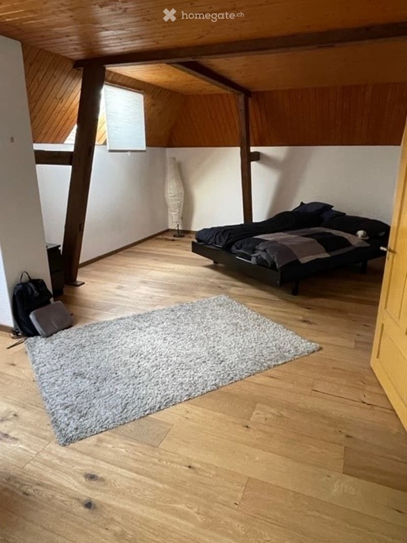 Hardwood floors, beams on the ceiling, bed, pillow, blankets, rug, a grey bag, and a white floor lamp