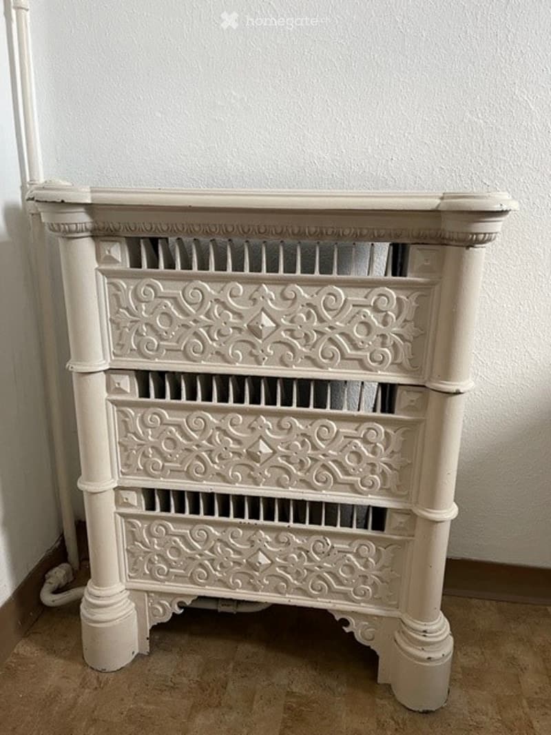 A vintage-style white radiator with ornate decorative patterns on its surface, mounted on a wall in a room with tiled flooring.
