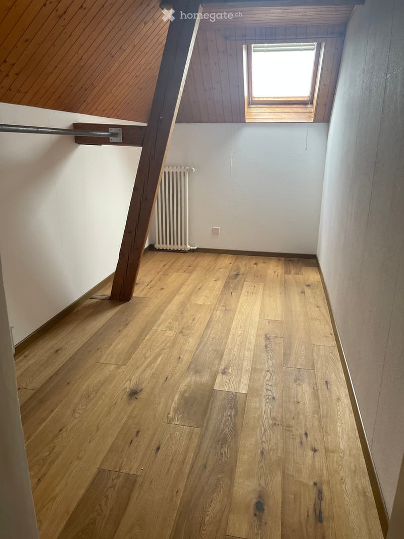 Empty attic room, wooden flooring, small window, wooden beams, white walls, radiator