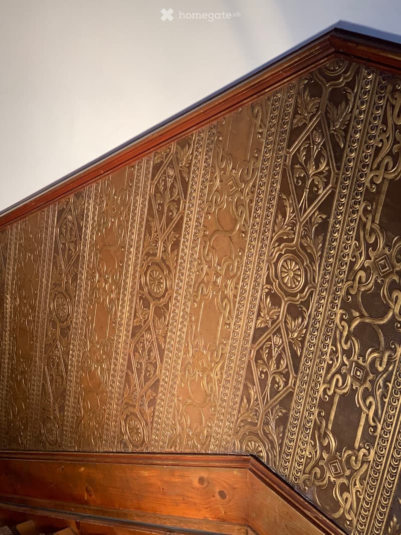 wall with intricate gold patterned wallpaper, wooden baseboard