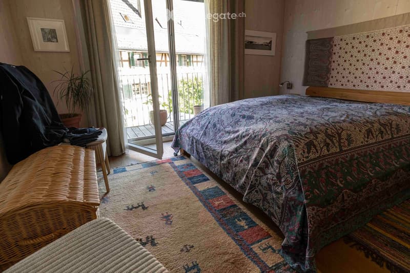 Cozy bedroom with a patterned bedspread, wicker chair, colorful rug, and large window leading to a balcony.