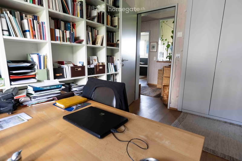 A well-organized home office with a wooden desk, laptop, chair, bookshelf filled with books, and a small plant in the background.