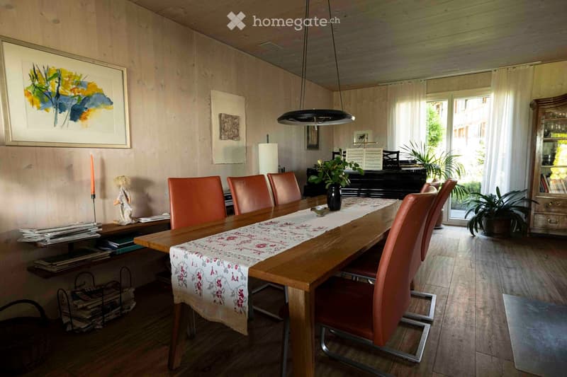 Dining room with a long wooden table, six orange chairs, a floral table runner, a black piano, a wooden cabinet, and potted plants. The room features wooden flooring, large windows with white curtains, and artwork on the walls.