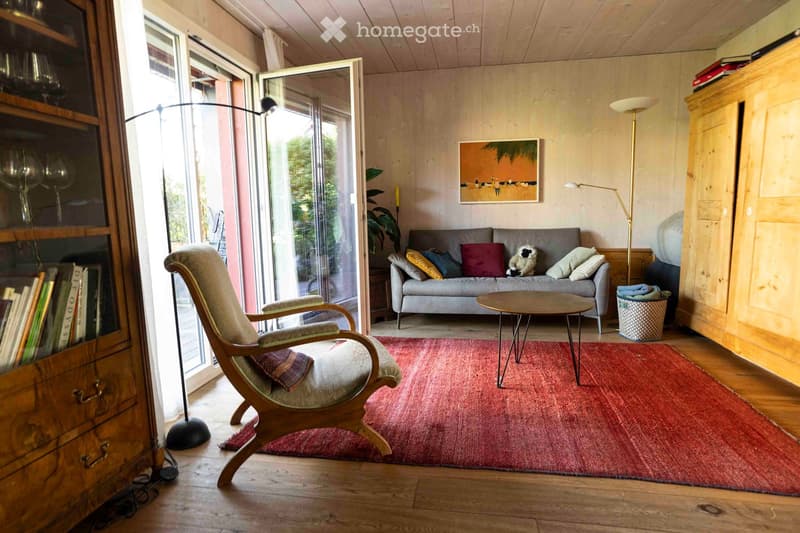 Bright living room with wooden floors, a red rug, a grey sofa with colorful cushions, a wooden armchair, a small coffee table, and a floor lamp. A bookshelf and a picture frame are visible on the walls.
