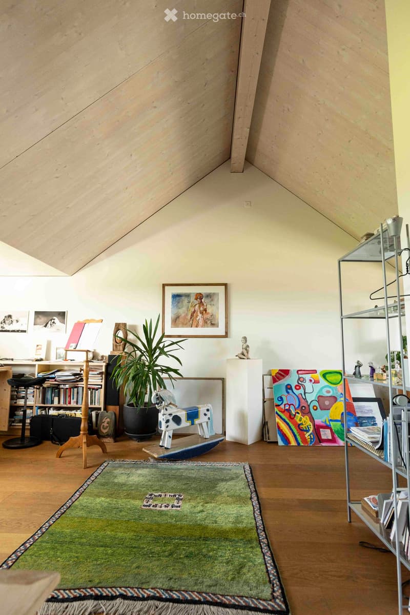 A cozy, well-lit attic room with wooden floors, a slanted ceiling, and various furnishings including a green rug, bookshelf, plant, and artwork.