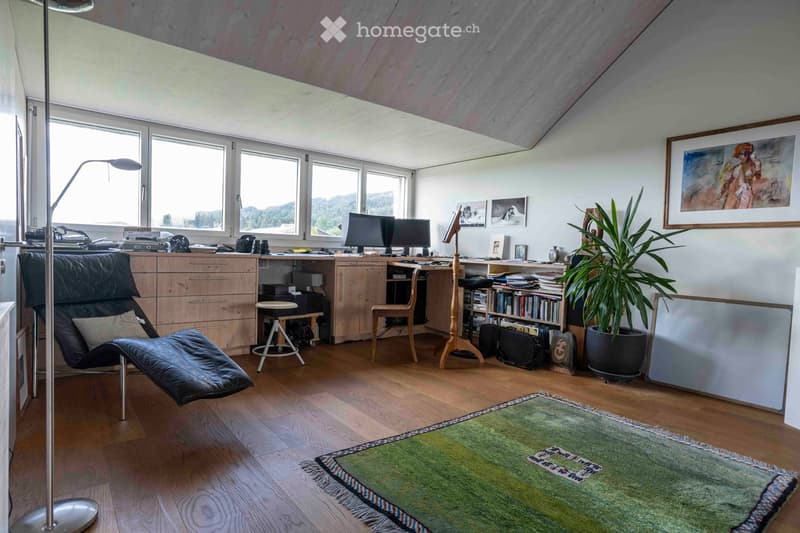A well-lit room with wooden flooring, large windows, a desk with two monitors, a black leather chair, a small stool, a potted plant, and artwork on the walls.