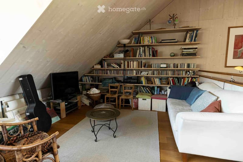 Cozy attic living room with slanted wooden ceilings, white sofa, coffee table, bookshelf filled with books, and a guitar case.