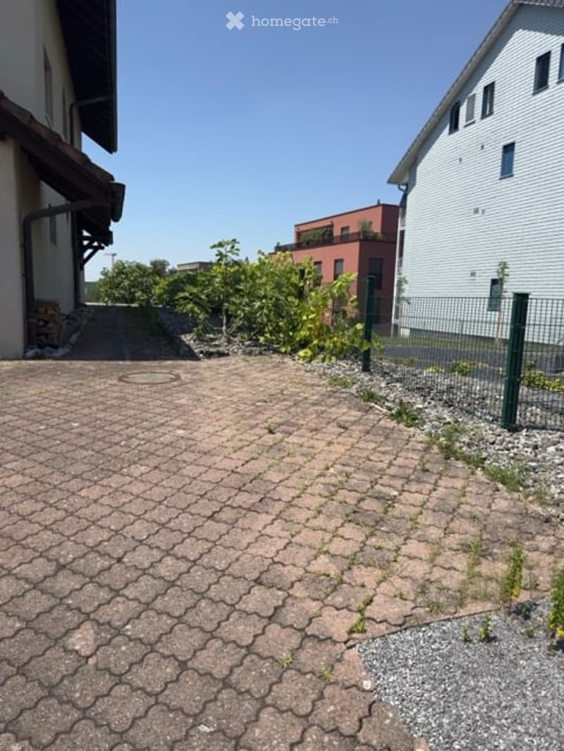 A paved outdoor area with a brick-patterned ground, surrounded by a green metal fence. There are some small plants and shrubs near the fence, and a few buildings are visible in the background under a clear blue sky.