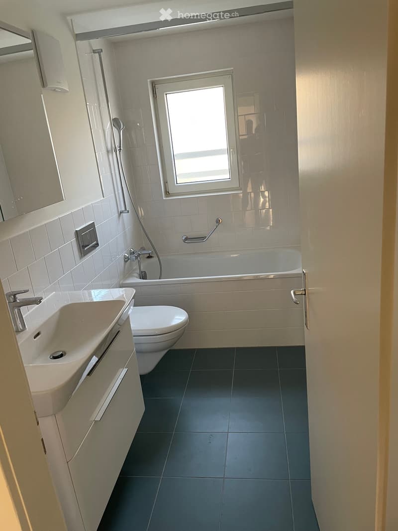 Modern bathroom with white tiles, a sink, toilet, bathtub, and a window above the bathtub. Features a sleek design with a dark tiled floor and a mirror above the sink.