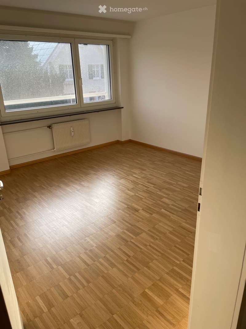 Empty room with light wooden flooring, white walls, large windows, and a radiator under the windows.