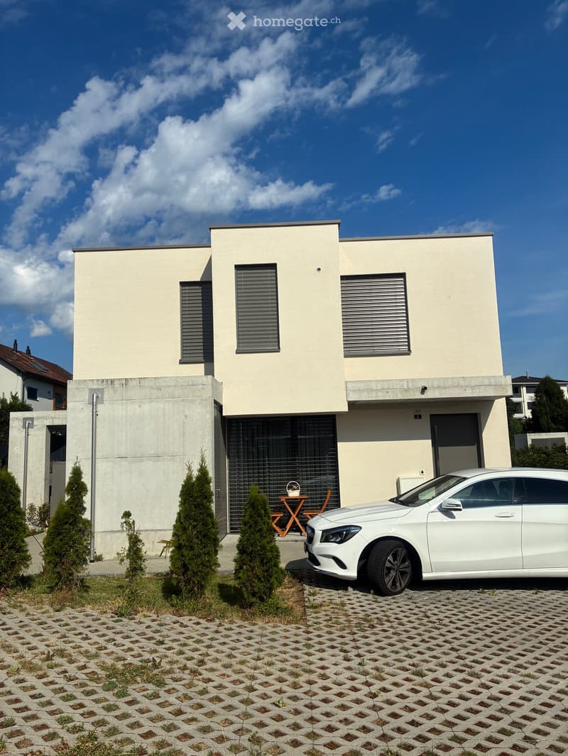 Modern two-story house with light beige facade, grey-framed windows, garage, and a small outdoor seating area with a table and chairs. A white car is parked in front.