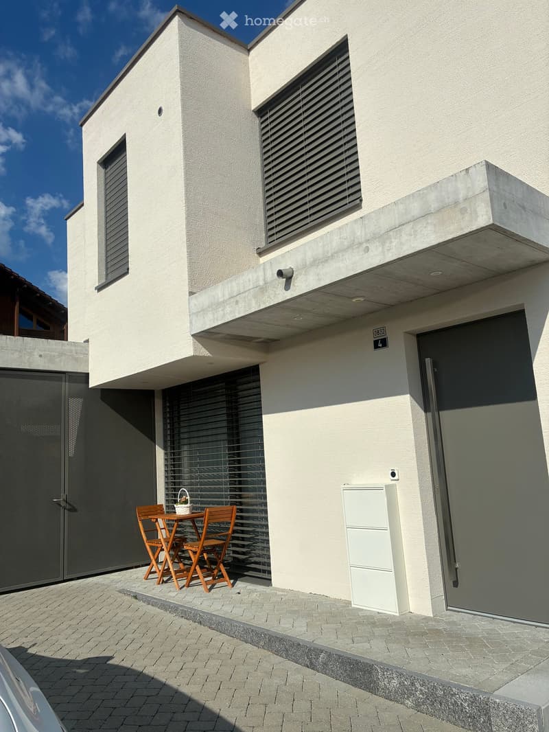 Modern house exterior with a small outdoor seating area featuring a wooden table and chairs, a white utility box, and a raised ground floor entrance.
