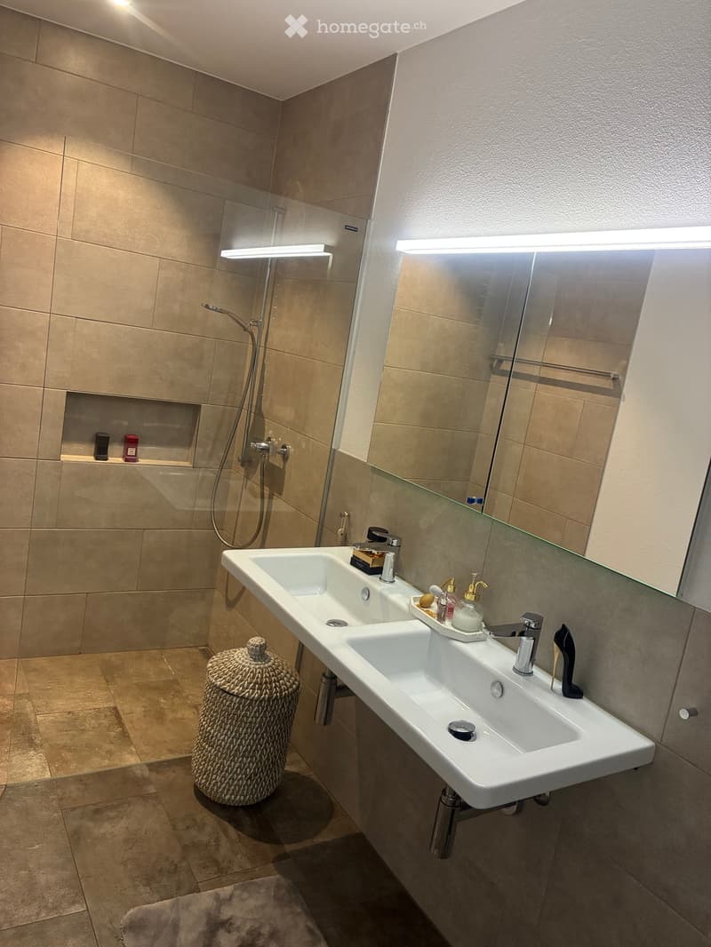 Modern bathroom with a double sink, large mirror, shower area, wicker basket, and various toiletries.