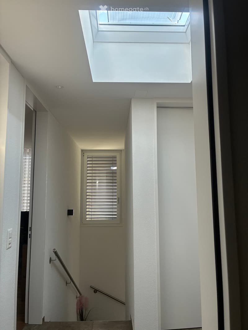 Modern hallway with white walls, a skylight at the top, a window with blinds, and a metal handrail on the left side.