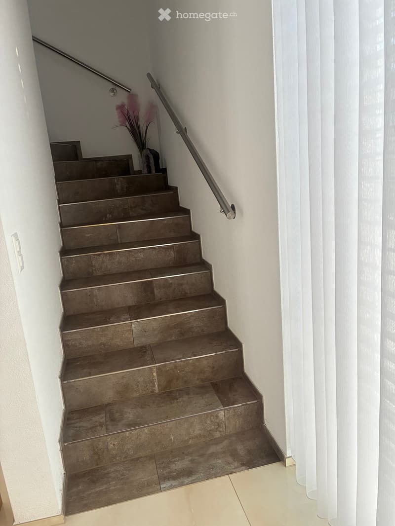 A modern staircase with gray tiled steps and a metal handrail, leading upwards. The walls are white, and a sheer white curtain is visible on the right side.