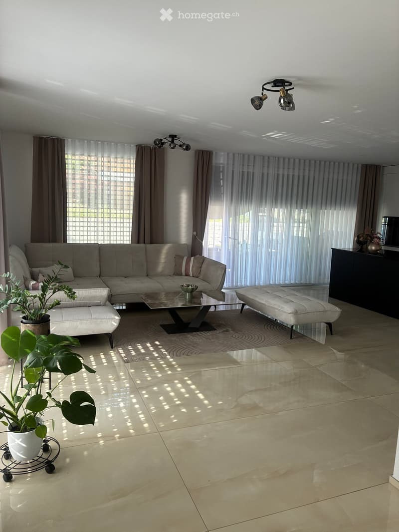 Modern living room with large windows, beige sofa, glass coffee table, white ottoman, potted plant, and a dark cabinet.