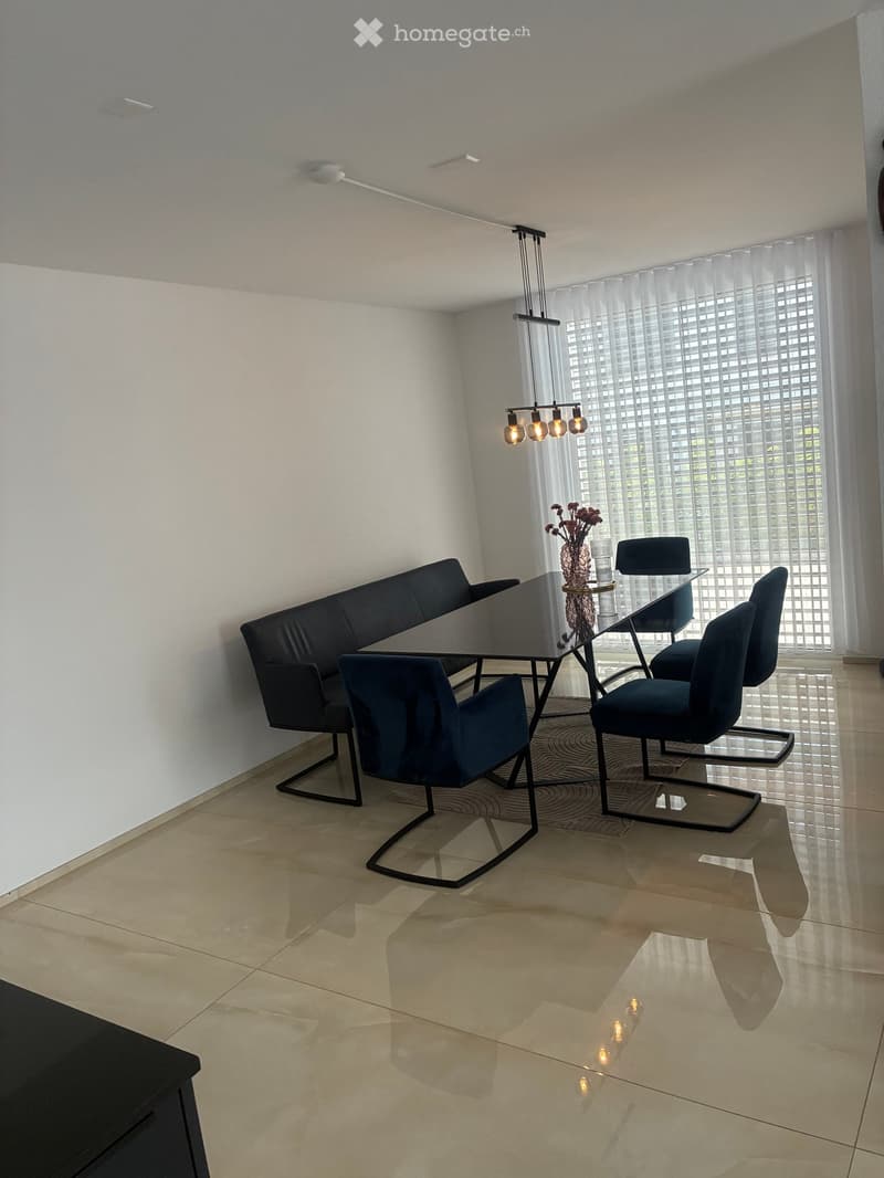 Modern dining area with a black sofa, glass dining table, four chairs, and a vase with flowers on the table. Large windows with blinds and a hanging light fixture are visible.