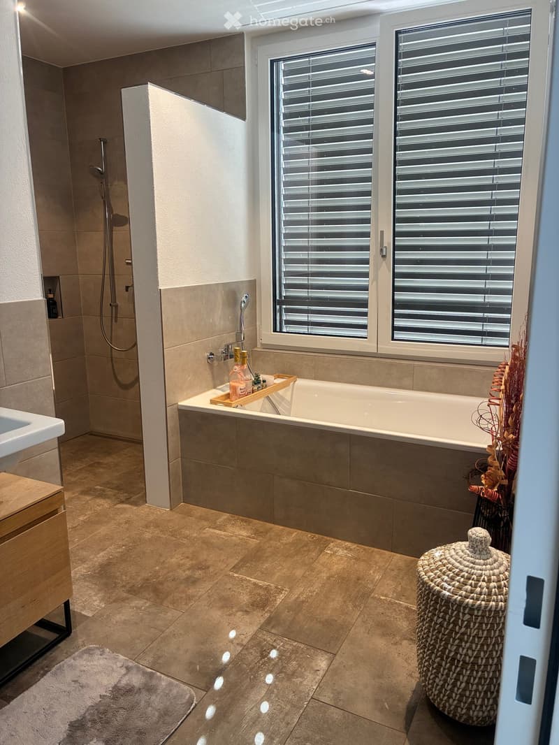 Modern bathroom with a bathtub, shower, sink, wooden tray with toiletries, wicker basket, and a soft rug on the tiled floor.