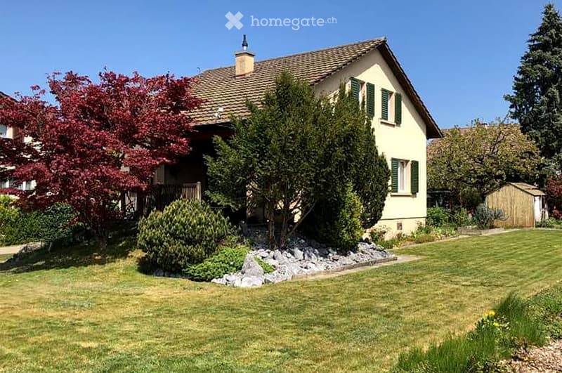 Two-story house with a tiled roof, green shutters, and a well-maintained garden with trees and shrubs.