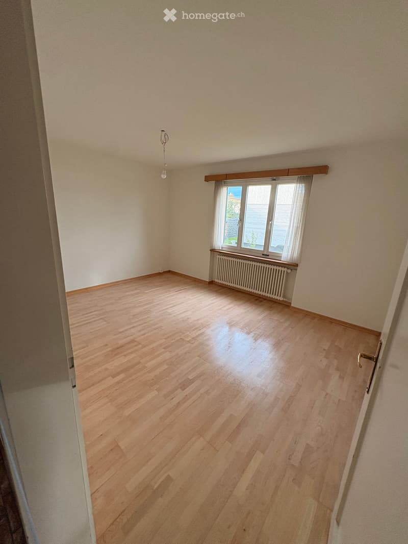 Empty room with wooden flooring, white walls, a window with white blinds, and a radiator below the window. The room has a simple, clean design with no furniture or decorations.