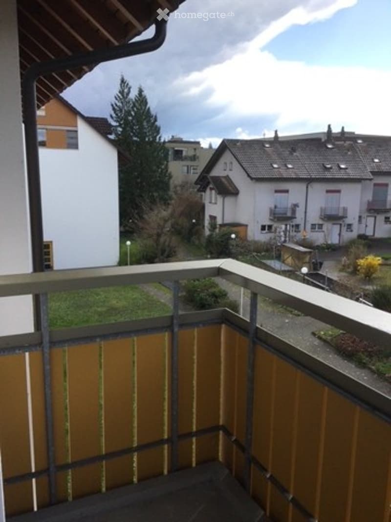 Balcony with metal railing, overlooking a residential area with houses, trees, and a clear sky.