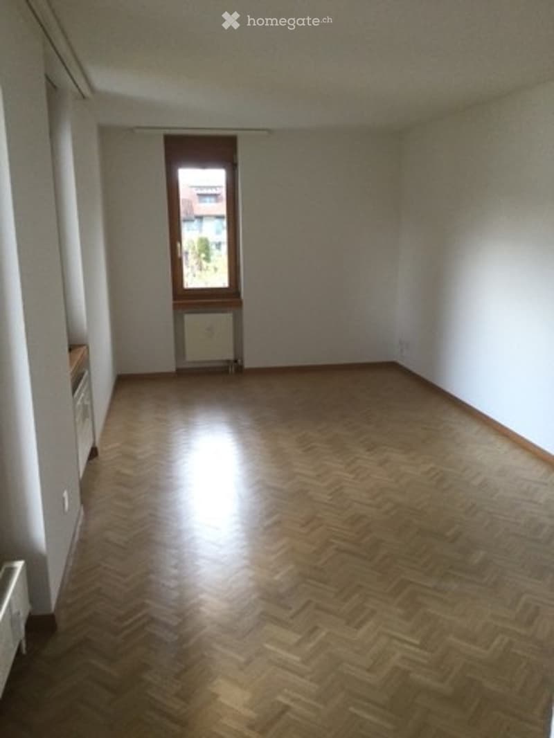 Empty room with wooden parquet flooring, white walls, and a single window with brown frame. No furniture or equipment present.