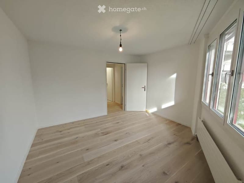 Empty room with light wooden flooring, white walls, and large windows allowing natural light. A single door leads to another room.