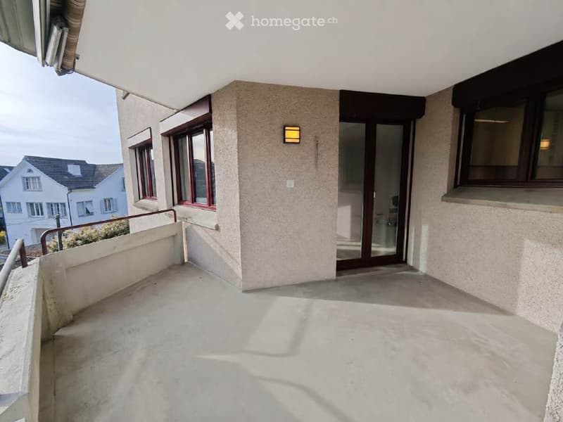 Balcony with concrete floor, metal railing, and glass door leading to interior. View of neighboring houses and greenery.