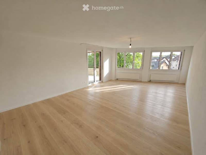 Empty room with light wooden flooring, white walls, large windows, and a sliding glass door leading to a balcony.