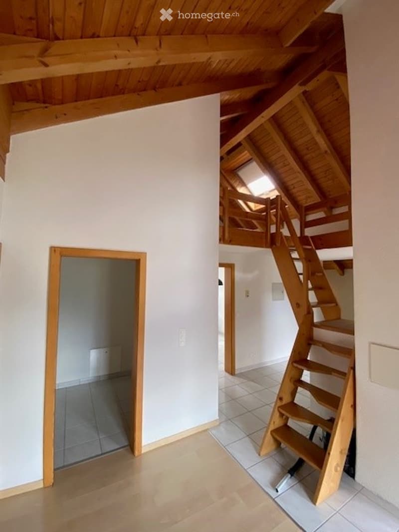 Bright, open-plan space with wooden floors, white walls, and exposed wooden beams. Features a staircase leading to an upper level with a small landing and a window.
