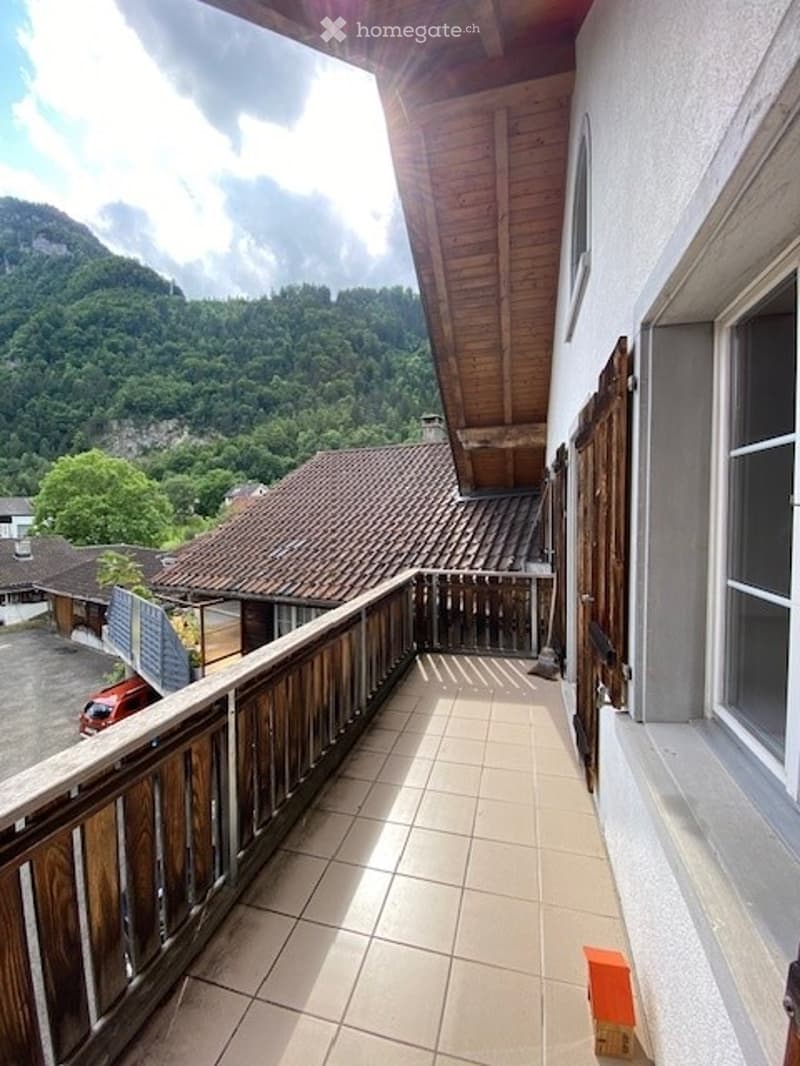 Balcony with wooden railing, tiled floor, overlooking a scenic mountain landscape with houses and trees.