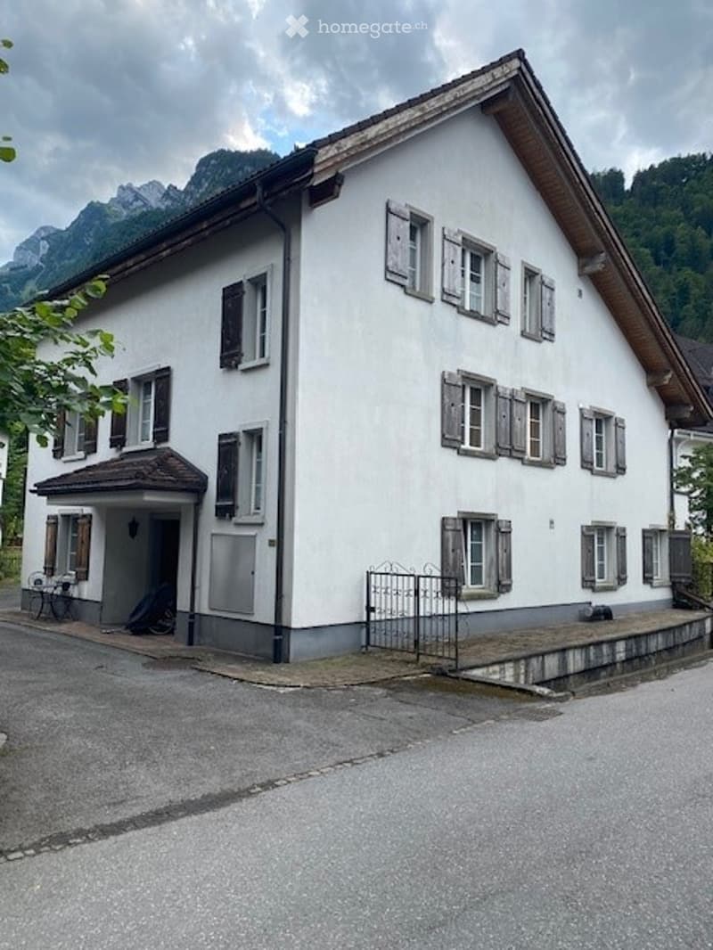 A two-story white house with multiple windows and shutters, situated in a mountainous area with a small porch and outdoor seating area.