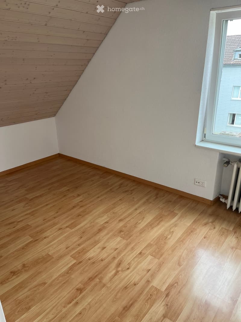 Empty room with wooden flooring and white walls, featuring a slanted wooden ceiling and a window with a view of neighboring buildings.