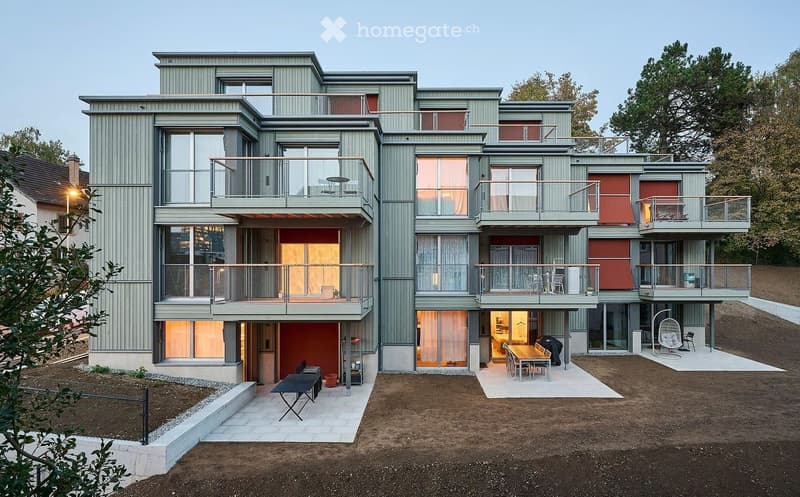 Modern, multi-story residential building with grey siding, large windows, and multiple balconies. The ground level features outdoor seating areas, a patio, and a small garden space.