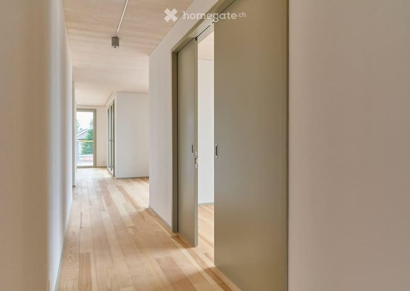 Modern hallway with light wooden flooring, white walls, and recessed lighting. Features several closed doors and an open doorway leading to another area.