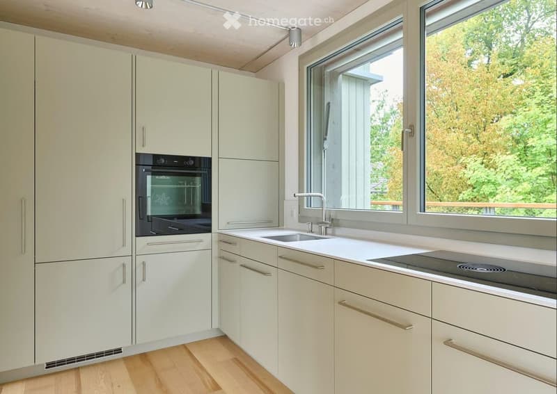 Modern kitchen with light-colored cabinets, a stainless steel oven, a sink with a modern faucet, and a stovetop, all set against a backdrop of large windows showing greenery outside.