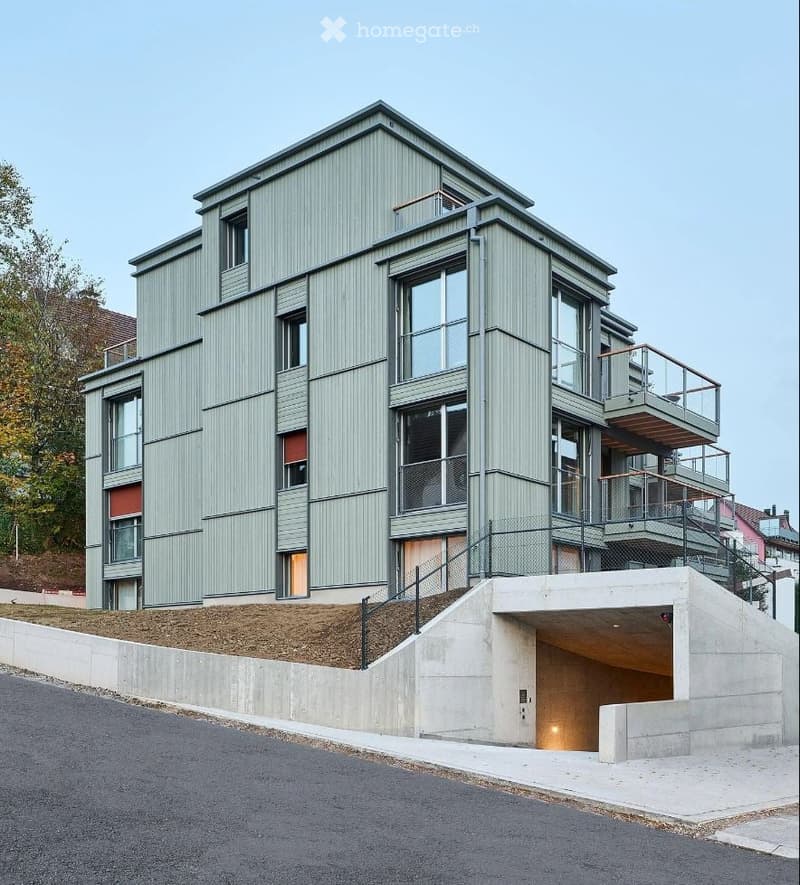 Modern three-story building with green siding, large windows, balconies, and a raised ground floor with a garage entrance.