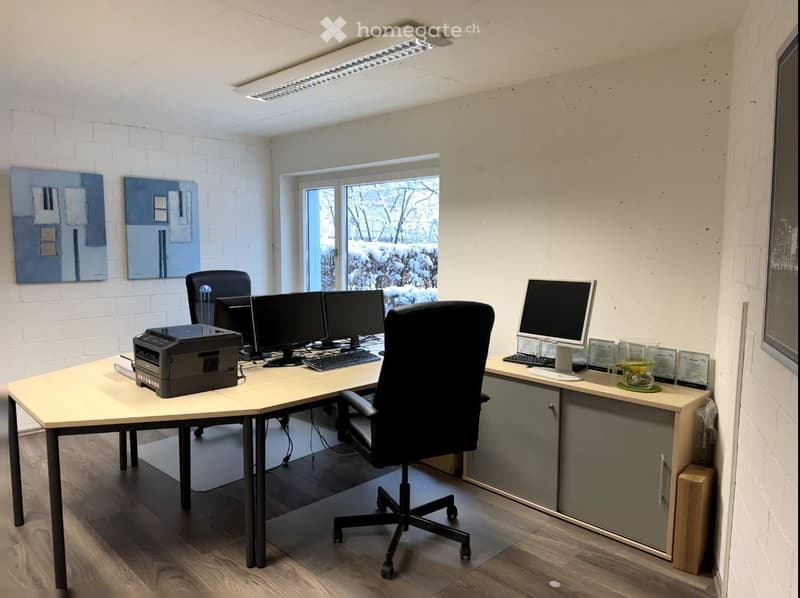 Modern office space with a large L-shaped desk, multiple computer monitors, a printer, ergonomic chair, and artwork on the walls. The room has white brick walls, large windows, and a sleek, contemporary design.