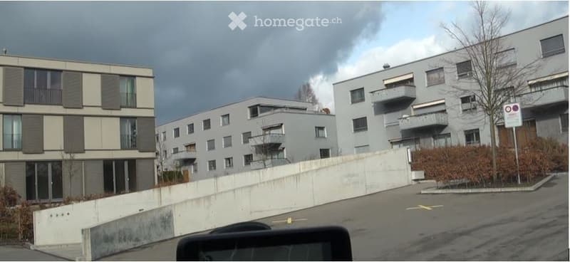 Modern multi-story residential buildings with balconies and large windows, surrounded by a paved area and low hedges.