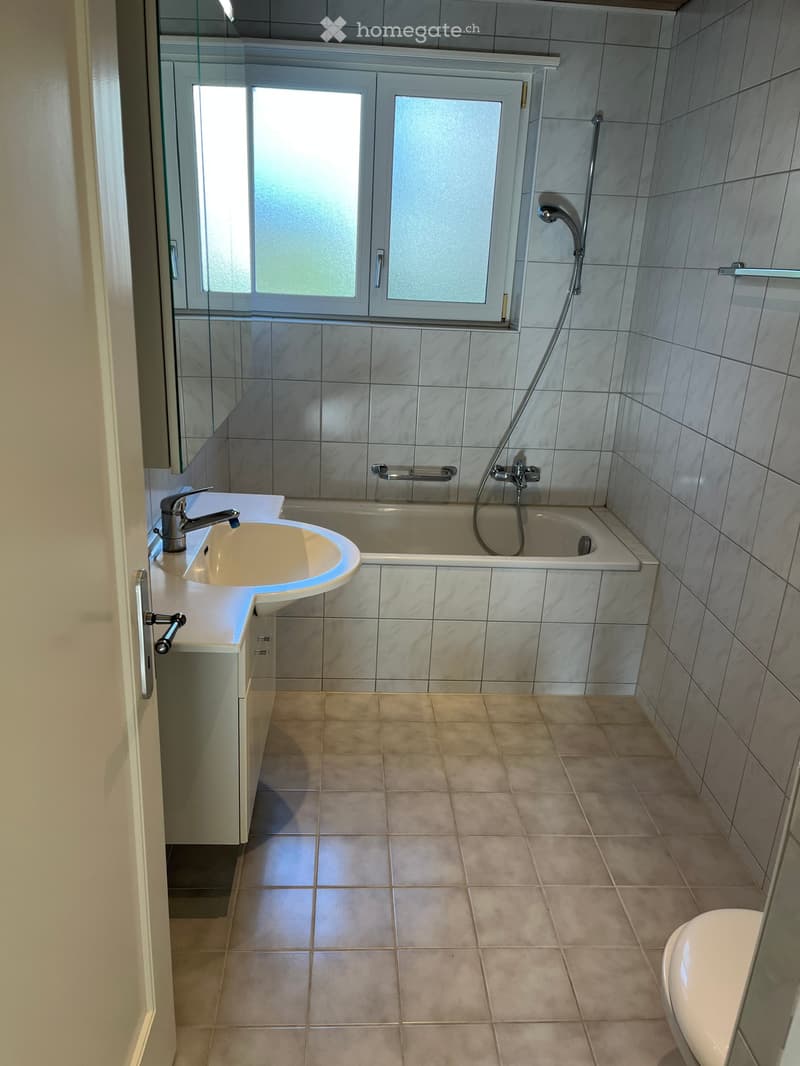 Modern bathroom with a white sink, bathtub, and toilet. Features marble tiles on walls and floor, a window with frosted glass, and a chrome showerhead.