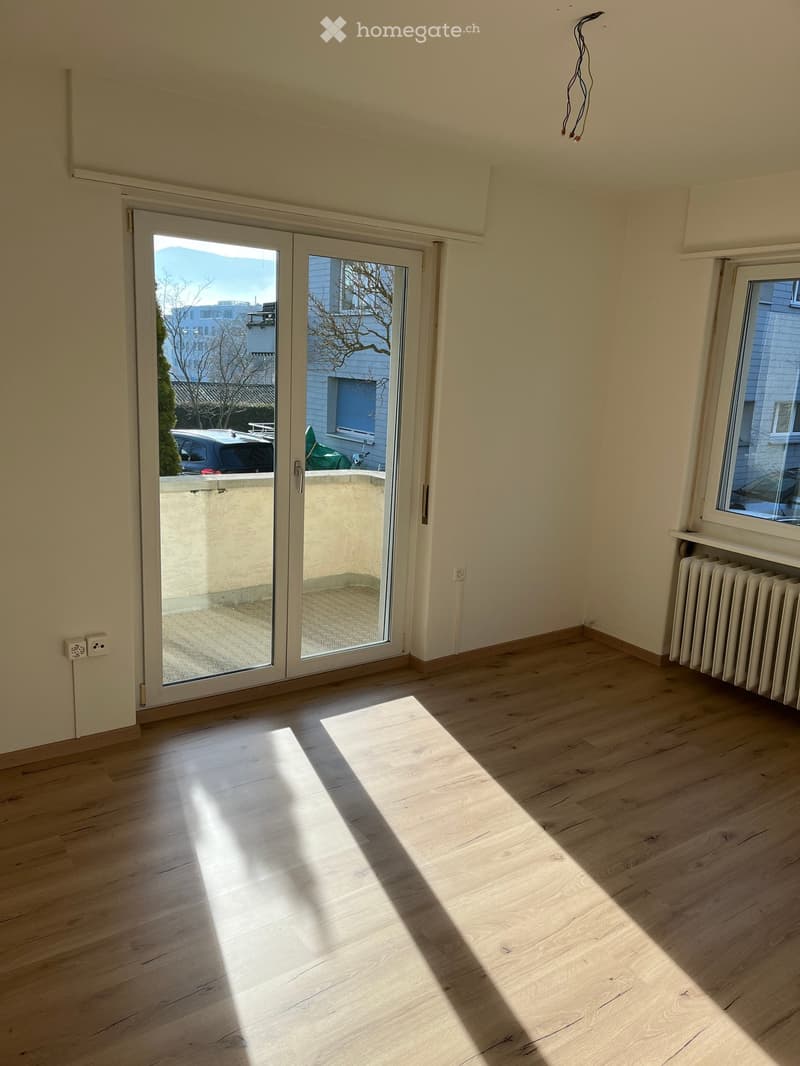 Empty room with light wooden flooring, large sliding glass doors leading to a balcony, and a radiator on one wall.