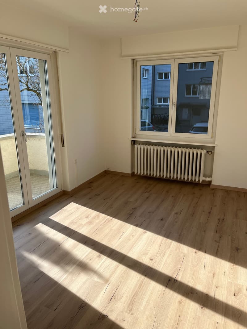 Empty room with light wooden flooring, white walls, large windows, and a radiator. Sunlight streams in from the windows, casting shadows on the floor.