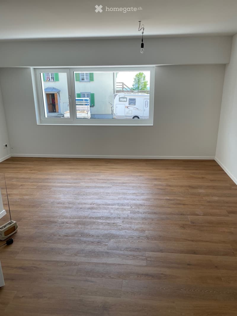 Empty room with wooden flooring and large windows, showing a view of a residential area with a camper van outside.