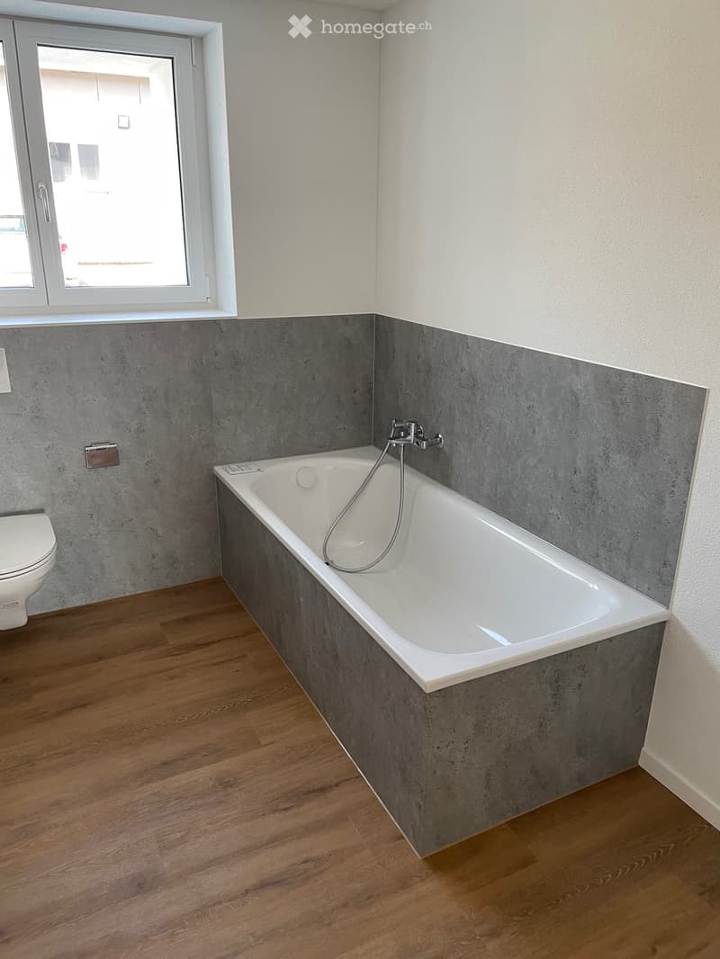 Modern bathroom with a white bathtub, grey tiled walls, wooden flooring, and a toilet. Features a large window with a view outside.