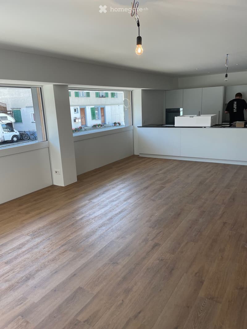Empty room with wooden flooring, white walls, and large windows offering a view of the street. The room is unfurnished, with only a counter and cabinets visible in the background.