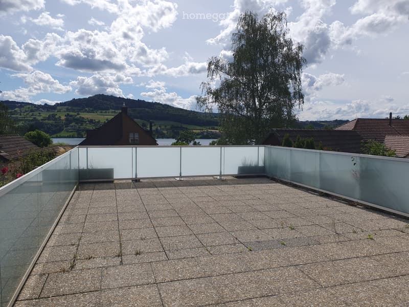 Modern rooftop terrace with a glass railing, tiled surface, and scenic views of a lake and surrounding hills.