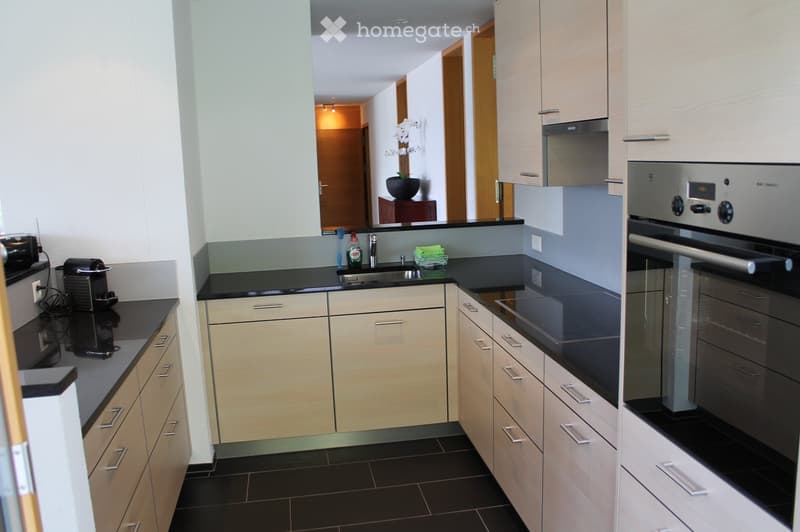 Modern kitchen with light wood cabinets, black countertops, and stainless steel appliances including an oven and coffee maker. Features a sink with a faucet and a small green towel on the counter.